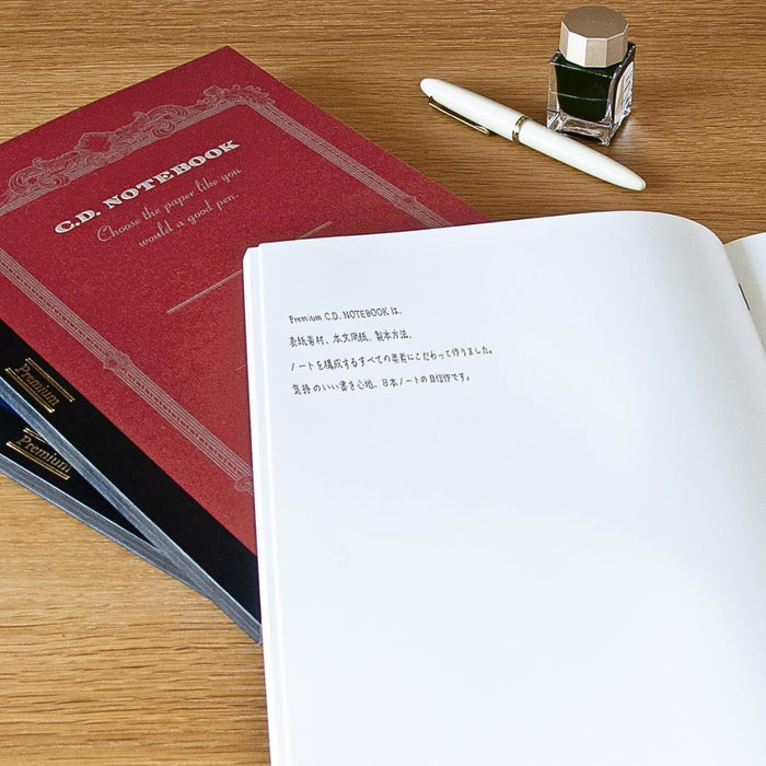 Apica Premium C.D Notebooks A4 Plain on a wooden desk with an open notebook and writing tools.