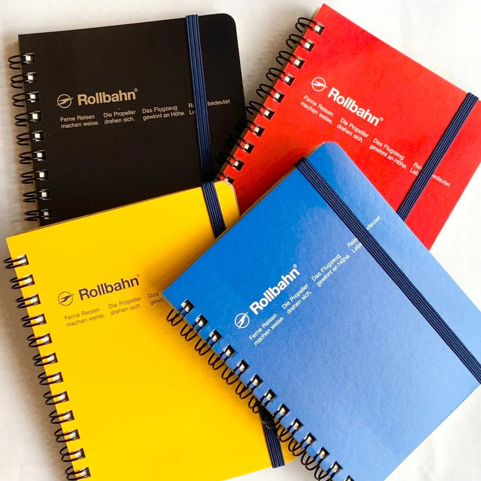 A flat lay of four colourful Rollbahn spiral notebooks in black, yellow, red, and blue, each secured with a navy elastic band.