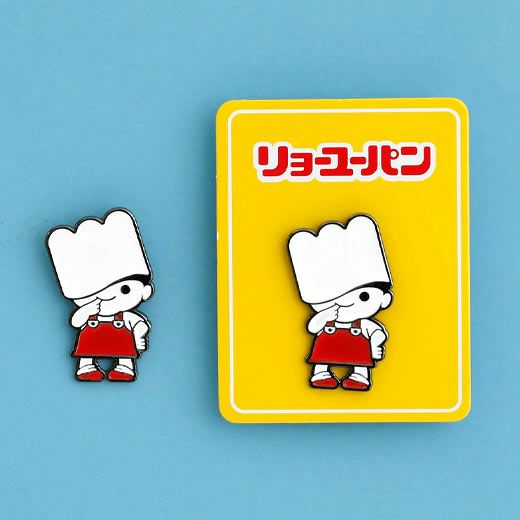 Cartoon chef enamel pin shown next to its bright yellow backing card with Japanese text, on a blue background.