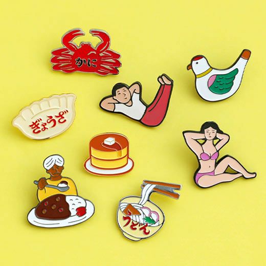 Collection of quirky enamel pins on yellow background, including bird, pancake, crab, gyoza, curry, ramen, and two stretching figures.