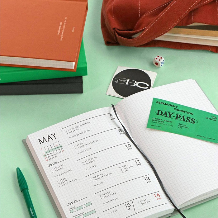 The Journal Shop - An orange Iconic Souvenir B6 Grid Notebook beside an open notebook with schedule and pen.