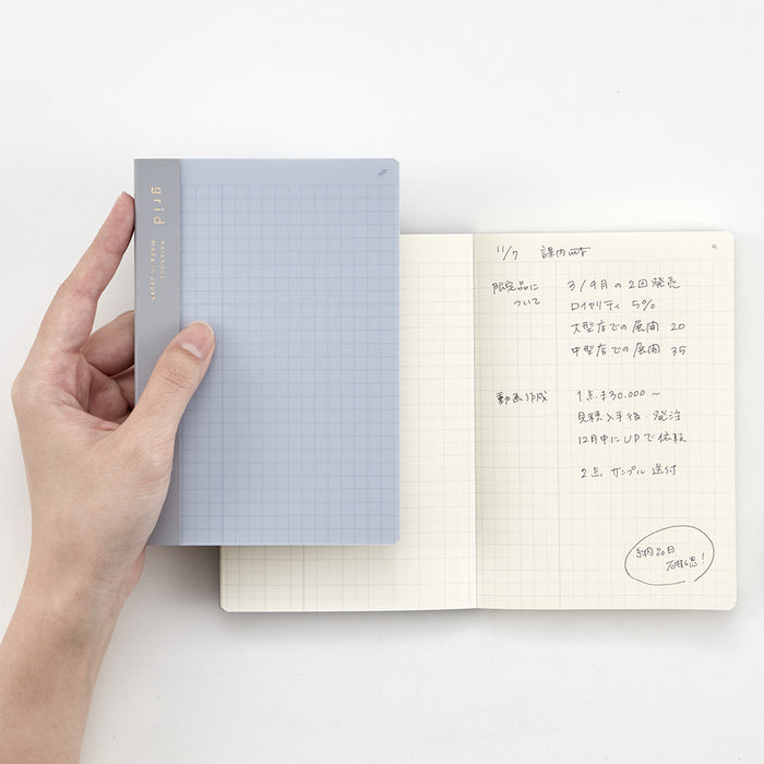 Hand holding the A6 blue Daigo Isshoni Number Notebook above an open sketchbook.
