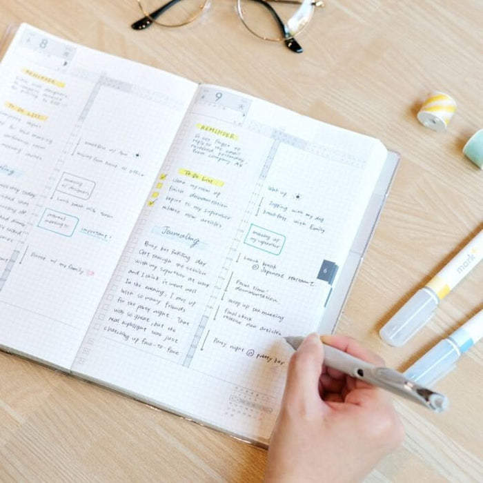 Person writing in Kokuyo Jibun Techo DAYs 2026 A5 Slim Grey edition at a desk with pen.