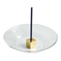 Brass cube incense holder centred on a round glass plate holding a single upright incense stick.
