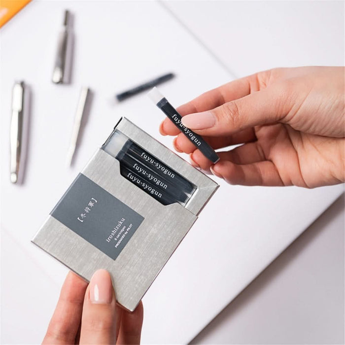 Hand holding a Pilot Iroshizuku Fuyu-syogun fountain pen refill ink cartridge with box on desk.