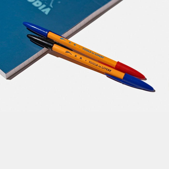 Tools To Live By dual-tip ballpoint pen in blue and orange placed on a notebook.