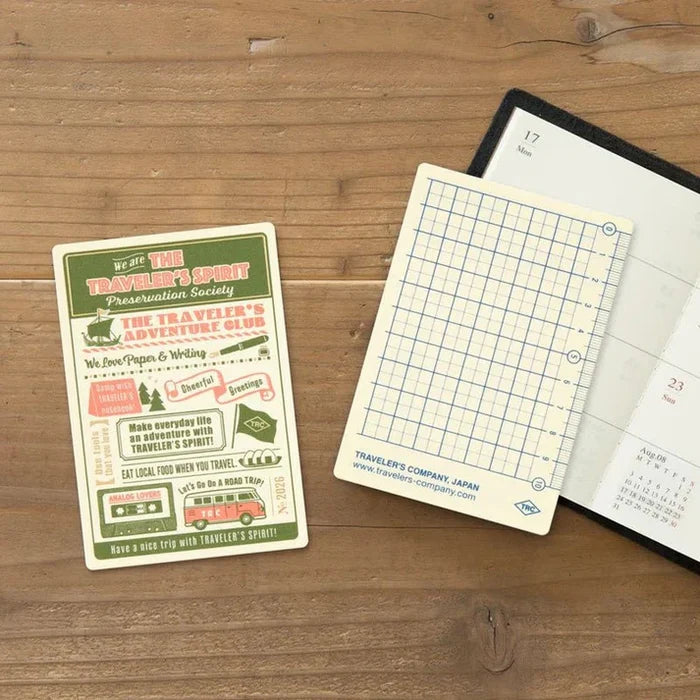 Traveler’s Notebook Passport Size Plastic Sheet 2026 Edition placed on a wooden desk beside an open notebook.