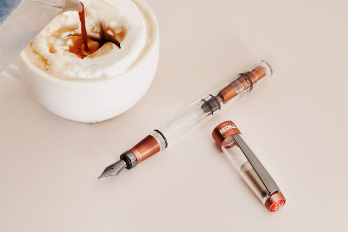TWSBI Diamond 580 fountain pen with copper aluminium accents placed beside a cappuccino as coffee is being poured into the frothy milk.