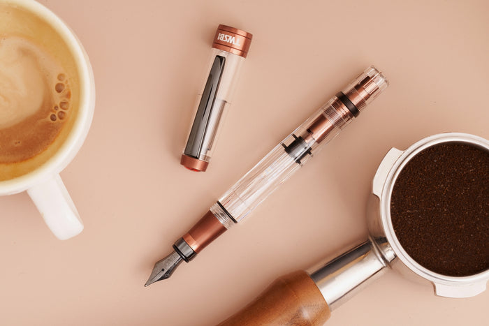 TWSBI Diamond 580 fountain pen with copper aluminium accents placed beside a cup of coffee and a portafilter filled with ground coffee on a beige surface.